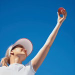 Woman reaching overhead to catch a red soft PU cricket ball under a clear blue sky.