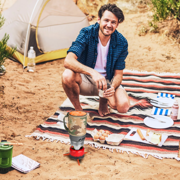 Camper using a portable camping stove with insulated mug while preparing food at a campsite, ideal for outdoor cooking, camping, and hiking.