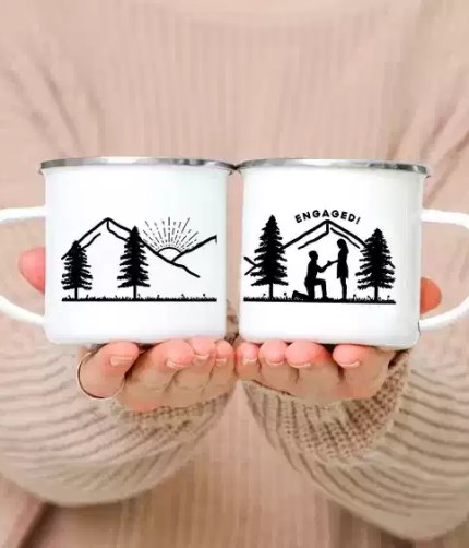 Two personalized engaged couple camp mugs with a creative woods print sitting on a wooden log.