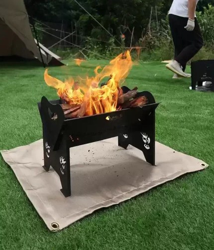 Portable fire blanket spread out on green grass under a burning metal fire pit to prevent scorching.