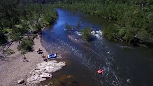 Aerial view of a serene river with kayakers and people relaxing on the shore, surrounded by lush greenery.