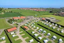 Aerial view of a well-organized campground with RVs and tents set on a lush green landscape, surrounded by fields and a distant sea view.