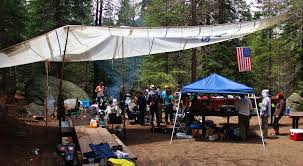 A group of people gathered at a campsite under a large tarp, with a blue tent nearby and a picnic area set up for cooking and socializing.