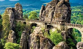 A scenic view of dramatic sandstone rock formations and a historic bridge in Saxon Switzerland National Park, Germany, surrounded by lush greenery and distant mountains.