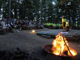 Campfire gathering with a lively audience watching a performance, surrounded by trees and a blazing fire.