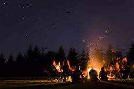 A group of people sitting around a campfire at night, with a starry sky above and the warm glow of the fire illuminating their faces.