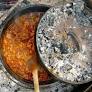 A pot of campfire chili simmering with a wooden spoon resting inside, partially covered by a lid.