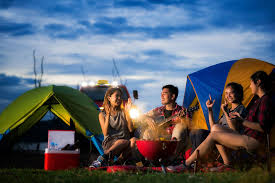 A group of four people sitting around a campfire in front of colorful tents, enjoying time together during a camping trip.