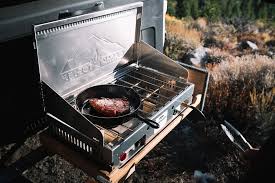 A portable camping stove set up with a frying pan cooking food outdoors, surrounded by nature.