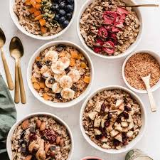 A variety of oatmeal bowls topped with fresh fruits, nuts, and seeds, presented on a light background.