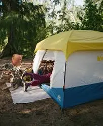A camping scene featuring a bright yellow and blue tent surrounded by outdoor chairs, a sandy area, and trees in the background.