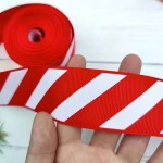 Hand holding a wide strip of red and white candy cane striped grosgrain ribbon next to a roll.