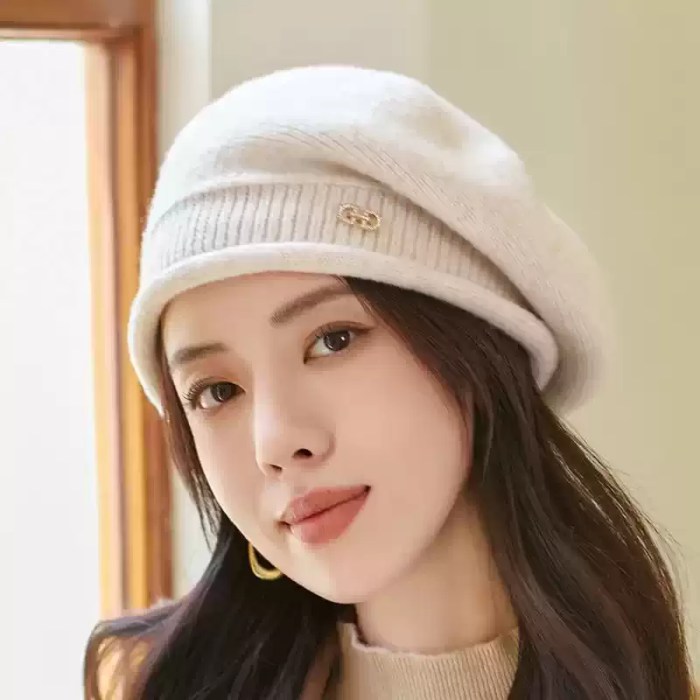 White Knitted Slouchy Beanie Hat - Interior Window View White knitted slouchy beanie hat with a textured rib knit band and metallic logo emblem photographed near an indoor window.
