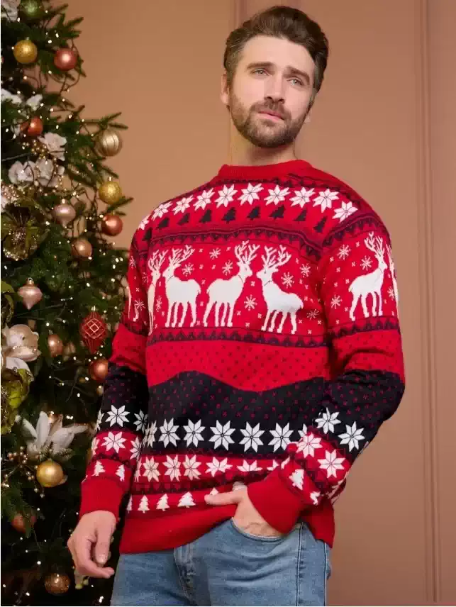 CHRISTMAS REINDEER SWEATER UK_2 A man and woman wearing matching red and black Christmas reindeer knit sweaters near a decorated tree.