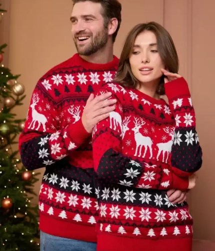 A man and woman wearing matching red and black Christmas reindeer knit sweaters near a decorated tree.