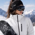 A woman wearing a black thermal ear warmer headband, white ski jacket, and ski goggles while holding a snowboard in a snowy mountain setting.