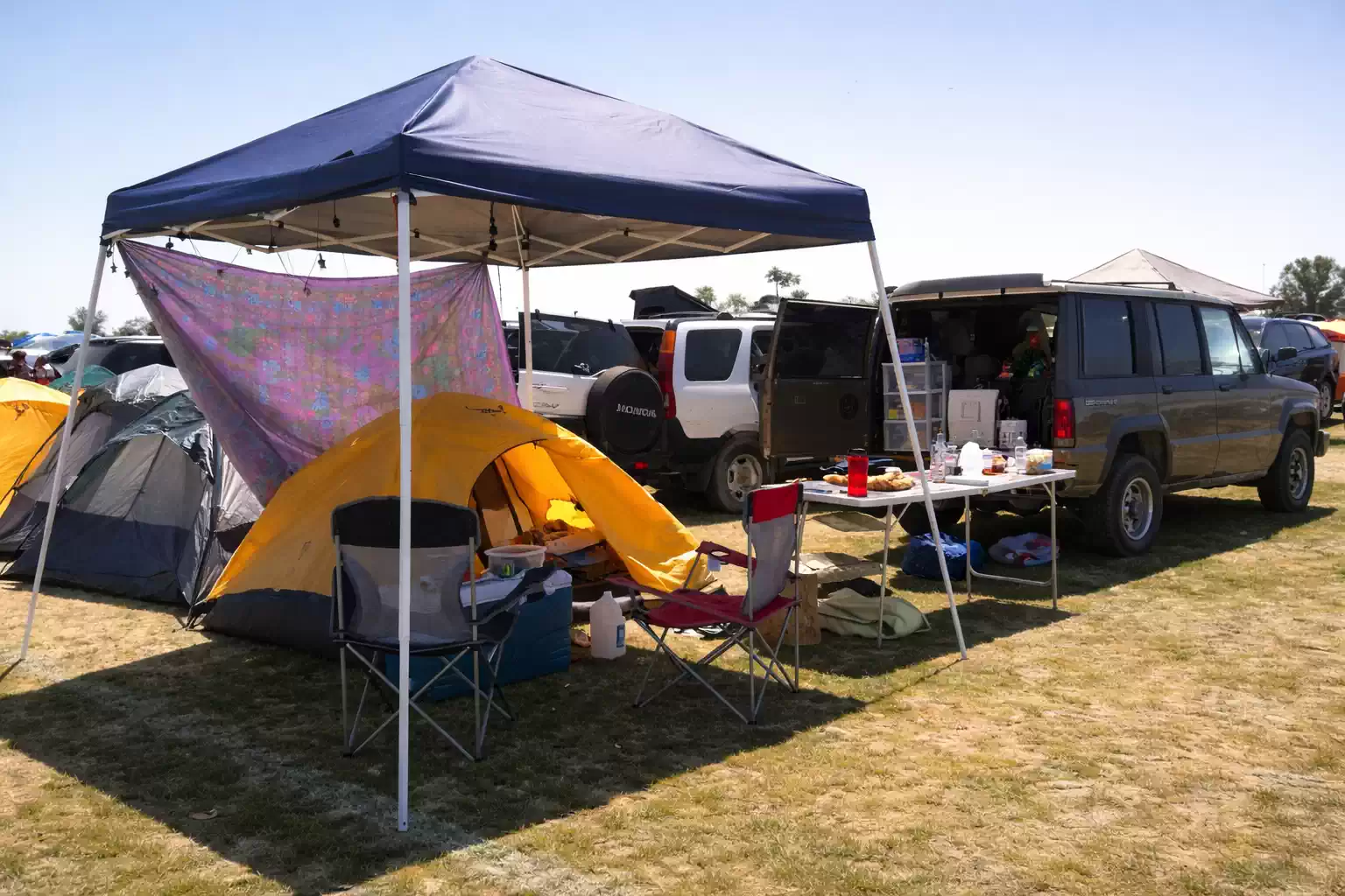 Daytime car camping setup with canopy tent, yellow camping tent, parked SUVs, folding chairs, table with cooking supplies, and shade cloth in an open campsite field